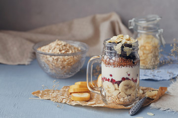 Layered dessert with muesli, yogurt, banana, linen seeds, sliced almonds and berries in glass mason jar on blue rustic background.