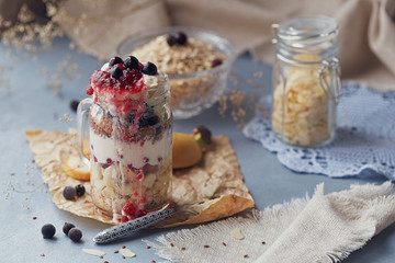 Layered dessert with muesli, yogurt, banana, linen seeds, sliced almonds and berries in glass mason jar on blue rustic background.