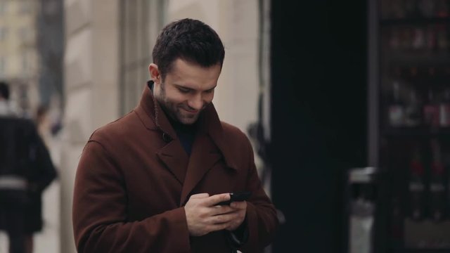 Young handsome man in a fashionable coat standing in the city center and using his phone for the Internet surfing, smiles. Modern lifestyle, technologies, social networks.