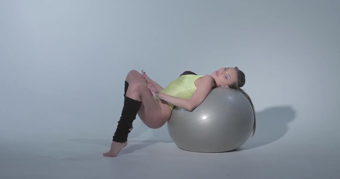 Beautiful girl in a yellow sports suit and black leggings is engaged in gymnastics on a gray rubber ball, swimsuit, beautiful skin, figure, barefoot, crouchs on the ball, jumps,  slog