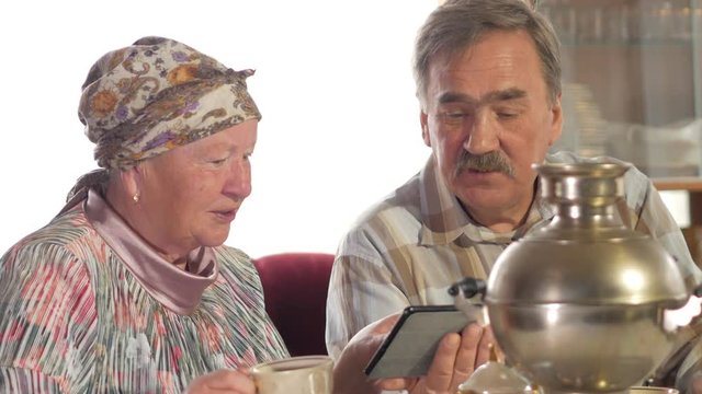 Elderly couple drink tea from a vintage Russian teapot samovar and watch a photo in the tablet. A man with a mustache and a woman discussing the image.