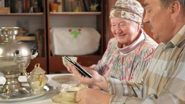 Elderly couple drink tea from a vintage Russian teapot samovar and watch a photo in the tablet. A man with a mustache and a woman discussing the image.