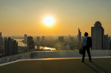 Fototapeta na wymiar Silhouette of businessman standing with carrying the business bag and looking the vision over the cityscape background at sunset time with lens flare,Business success concept