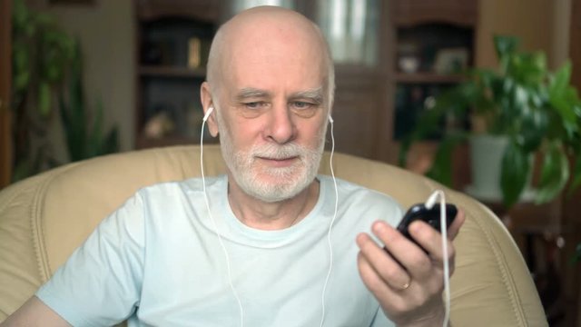 Good-looking senior man sitting in chair at home. Listening music on smartphone with ear-phones