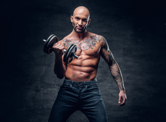 Obraz na płótnie Canvas Shirtless shaved head, muscular male with tattoos on his chest and arms holds dumbbell.