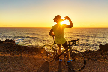 Athlete cyclist man drinking water after intensive biking training, enjoying sunset and ocean. Healthy active lifestyle sports fitness man resting on bike with sun flare.