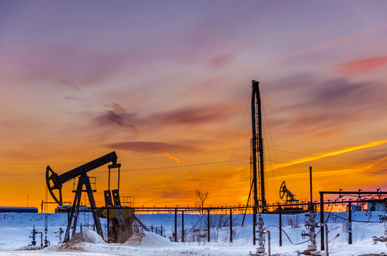 Pump jack, wellhead, pipeline and oil rig during sunset in the oilfield. Winter period. Oil and gas concept.