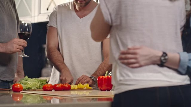 Tracking shot of men and women standing at kitchen counter and drinking wine while one of friends cooking vegetable salad