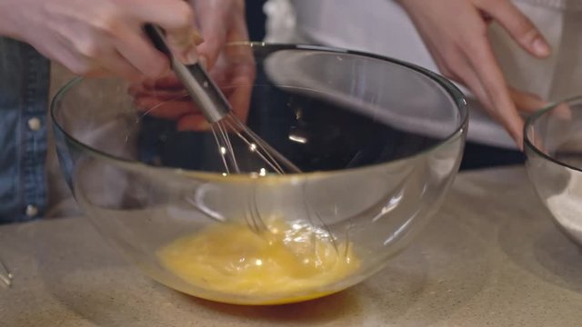 Closeup shot of female hands stirring eggs in glass bowl with whisk and hands of another woman adding flour
