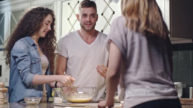 Tracking shot of friends cooking together in the kitchen: young woman whisking batter in glass bowl, man telling something to girls and they laughing