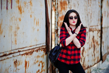 Fashion portrait girl with red lips wearing a red checkered shirt, sunglasses and backpack background rusty fence.