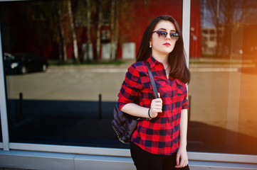 Fashion portrait girl with red lips wearing a red checkered shirt and backpack with sunglasses background large mirror windows.