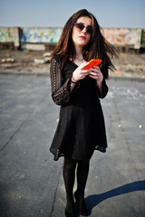 Portrait brunette girl with red lips and orange mobile phone at hands, wearing a black dress, sunglasses posed on the roof. Street fashion model.