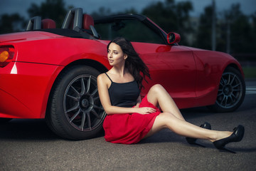 Cute young brunette woman sitting on the ground near the wheel of luxury red cabriolet car. - obrazy, fototapety, plakaty