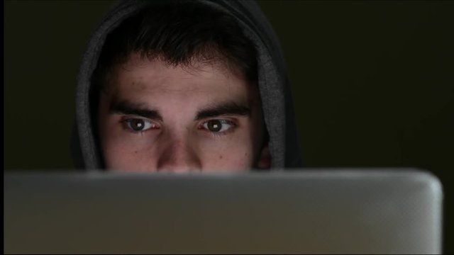 Cyber Criminal Using Laptop With Face Illuminated By Screen