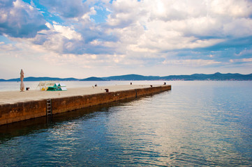 Fototapeta na wymiar A stone pier with metal ladders in the rippled sea water against a hill range in the distance and dramatic cloudy morning sky. Zadar, Croatia