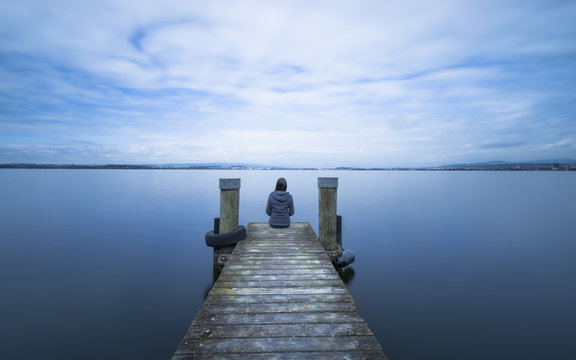 Overcast. Self reflection in magical world of fantasy. One woman sits on a wooden pier. Cloudy above the lake. Long exposure.