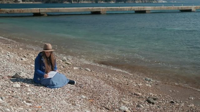 Young woman writing while sitting on beach. Brown-haired female in hat dressed in light jacket and long skirt rested comfortably on stones against background of blue sea and visible rocks. Cute lady