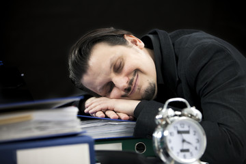 Obraz premium An employee is sleeping peacefully at his workdesk. There is an alarm clock on his table, too.