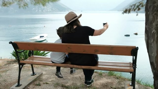Woman and boy sitting facing sea taking pictures on phone. Brunette dressed in hat, black jacket and jeans have good time with his son at beach on bench. Enchanted by beautiful scenic views and