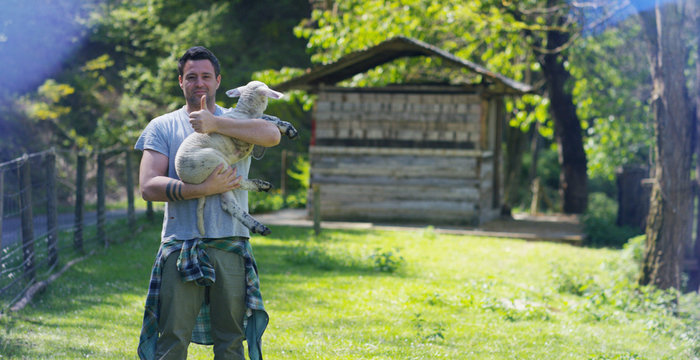 A young handsome and smiling farmer, holding a lamb young cub, has the experience to follow and care for live animals, on the background of nature and a barn, concept: ecology,livestock,bio,farming