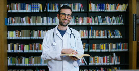 A young and beautiful doctor in a library smiling happy and holding books after doing a search and...