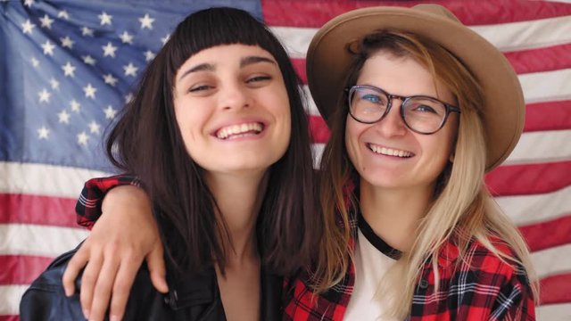 Two young students embrace and celebrate independence day in excitement and awe they laugh and giggle, hug they wear hipster clothing, hard core american jacket and flag hangs on the wall