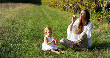 Fototapeta premium Super slow motion of young mother and baby 2 years old girl blowing air/soap bubbles in a meadow of vineyard on a sunny day in 4k 