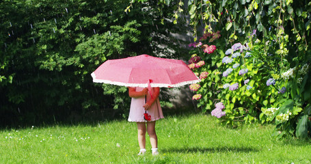 little girl in the garden playing with parents hiding under a red umbrella against splashing water and looking in camera . concept of security and saving of energy