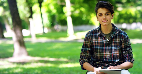 young student boy in the checked shirt use the tablet in a park in a moment of relaxation and enjoys his day