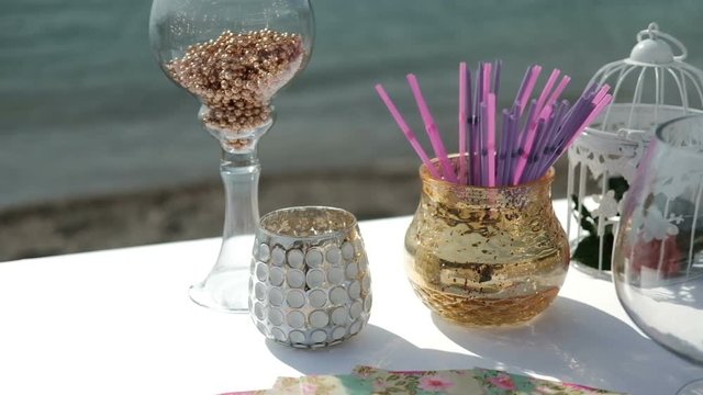 Glasses and vases on table by sand seashore. Transparent wineglasses with balls and shiny bowl with plastic tubules are felicitously located on white spacious desk in background of noisy faltering