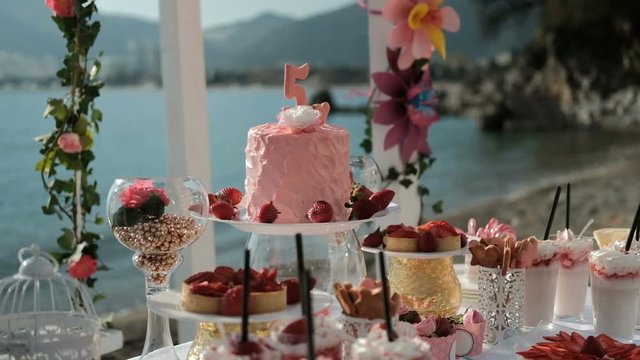 Cake, sweet dishes and flower compositions on table by sea. Wonderful banquet desk filled with lot of dulcet snacks and drinks in center of which rises huge pink pie with candle with figure of five