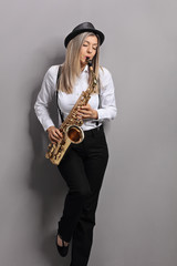 Fototapeta na wymiar Young woman playing saxophone and leaning against gray wall