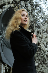 Girl with blond hair dressed in a black coat is standing under an umbrella