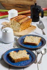 Coconut and rhubarb cake. White wooden table, dark blue plates, silver cutlery. 