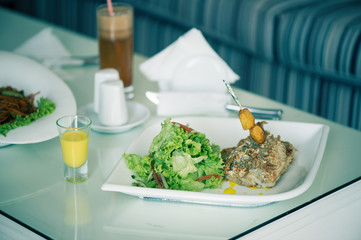 Fish with a salad in a plate is servered on a table with a glass of carrot juice. The female is having lunch.