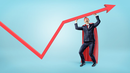 Fototapeta premium A businessman in a superman red cape supporting a red statistic arrow with his hands.