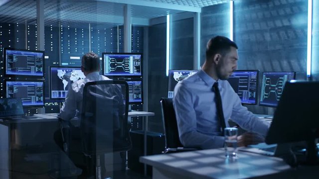 Two IT Professionals Working in a Monitoring Room with Multiple Displays Surrounding Them.System Control Room Has It's on Server Racks.  Shot on RED EPIC-W 8K Helium Cinema Camera.