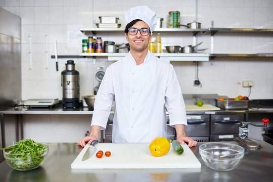 Happy young chef in uniform standing by workplace in the kitchen