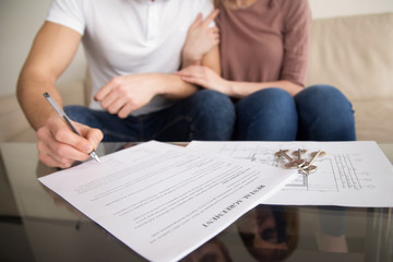 Close up of tenants signing rental agreement, renters couple sitting on couch, male hand with a pen putting signature, focus on document and keys, joint residential tenancy, lease contract - obrazy, fototapety, plakaty