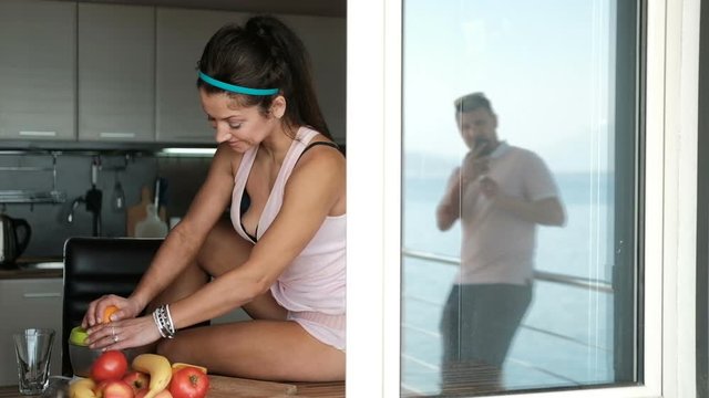 Young woman squeezes orange juice sitting by window in kitchen. Sweet brunette in pink body with bare legs coquettishly settled on table on which stand juicer and fruit. Fascinated by process of