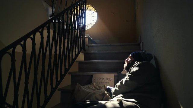 Homeless lying on the twilight stairs looks at the light coming from the window