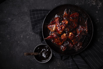 Chicken wings in thick barbecue sauce with garlic on black cast iron pan on dark rustic background.