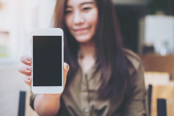 Mockup image of a beautiful woman holding and showing white mobile phone with blank black screen with smiley face in vintage wooden cafe
