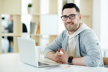 Portrait of modern middle-aged man smiling and looking at camera while working with laptop in sunlit office