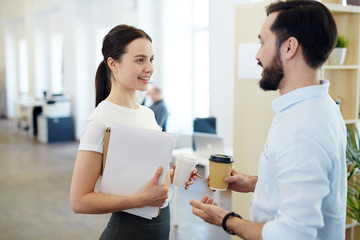 Portrait of smiling young woman talking to colleague during break in office, both holding paper coffee cups