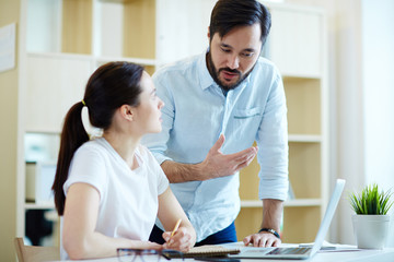 Portrait of adult Asian man leaning to help young woman at desk in office and discuss work