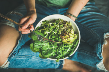 Green vegan breakfast meal in bowl with spinach, arugula, avocado, seeds and sprouts. Girl in jeans holding fork with knees and hands visible, top view. Clean eating, dieting, dieting food concept - obrazy, fototapety, plakaty