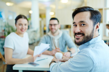 Team of cheerful business people smiling and looking at camera while meeting at table in modern office, enjoying work