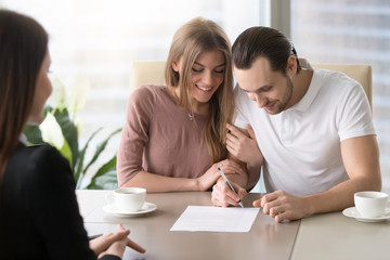 Smiling couple agreed to sign prenuptial contract, handsome man putting signature on document while sitting together with his wife, taking bank loan, health insurance, signing financial papers